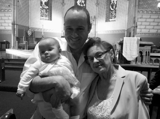 A family photo showing a man holding a baby alongside an older woman indoors